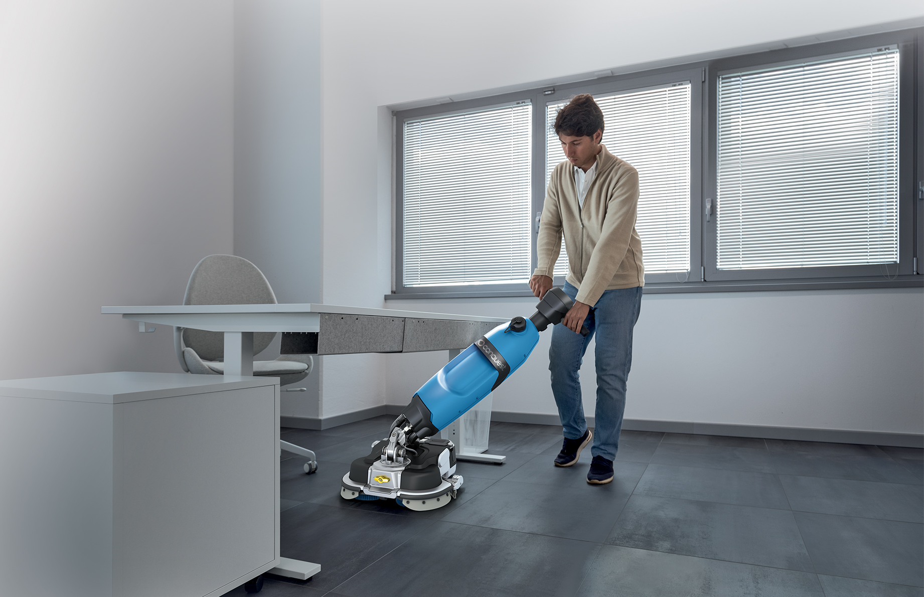Man operating a blue floor-polishing machine in an office, standing beside a desk and chair by large windows with blinds.