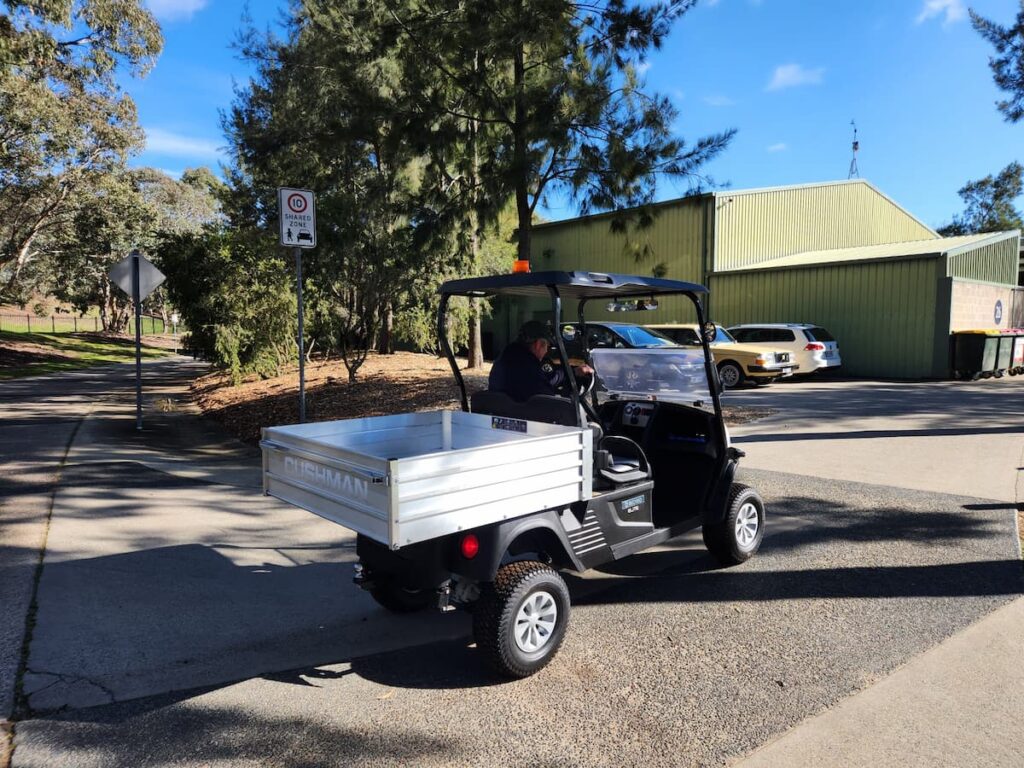 Small flatbed utility vehicle with a white cargo bed and an orange beacon parked in a sunny lot near a green warehouse; driver seated under the canopy.Small flatbed utility vehicle with a white cargo bed and an orange beacon parked in a sunny lot near a green warehouse; driver seated under the canopy.Small flatbed utility vehicle with a white cargo bed and an orange beacon parked in a sunny lot near a green warehouse; driver seated under the canopy.Small flatbed utility vehicle with a white cargo bed and an orange beacon parked in a sunny lot near a green warehouse; driver seated under the canopy.